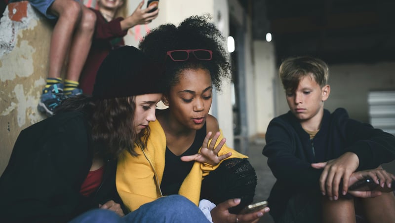 Two teenage girls are intently focused on a smartphone one of them is holding, while a teenage boy sits nearby looking down at his own phone. They are gathered in an urban, worn environment with another child visible in the background.
