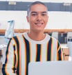 A smiling young adult with a shaved head and nose piercing looks directly at the camera while sitting at a desk with a laptop in a bright, modern indoor setting. They appear friendly and engaged.