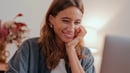 An adult woman with long hair smiles broadly and looks down at a screen, likely a laptop, with her hand resting under her chin. She appears happy and engaged in a bright indoor setting.