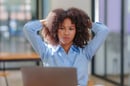 A teen with curly hair sits at a desk in front of a laptop, holding their hands behind their head, conveying a sense of stress or overwhelm.