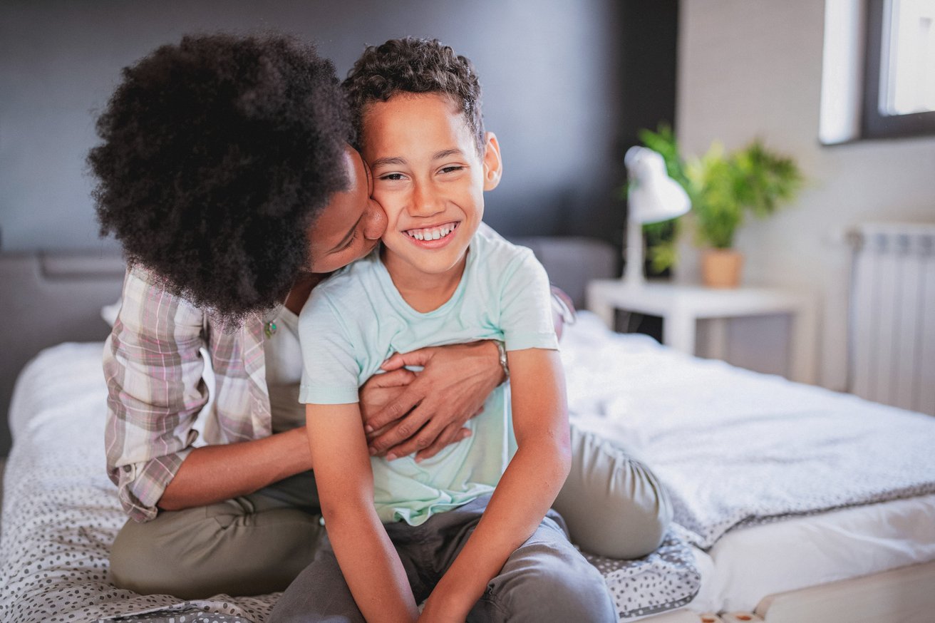 An adult is affectionately kissing a young child's cheek while sitting together on a bed. The child is smiling brightly at the camera, looking happy and secure.