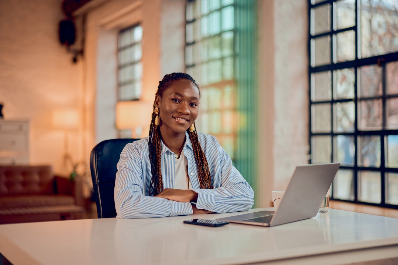 A smiling teenager sits confidently at a desk with a laptop and smartphone in a bright, modern office. She looks directly at the viewer with a friendly expression.