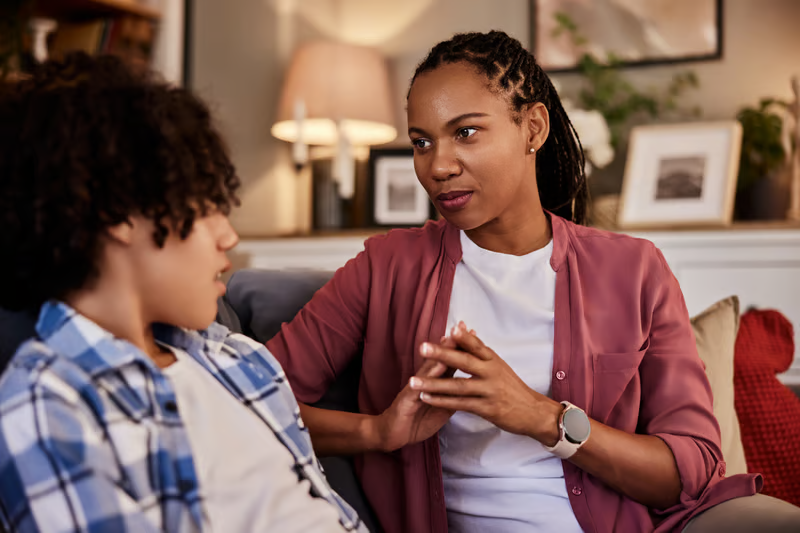 An adult woman sits on a living room sofa, listening attentively and speaking supportively with a teen beside her.