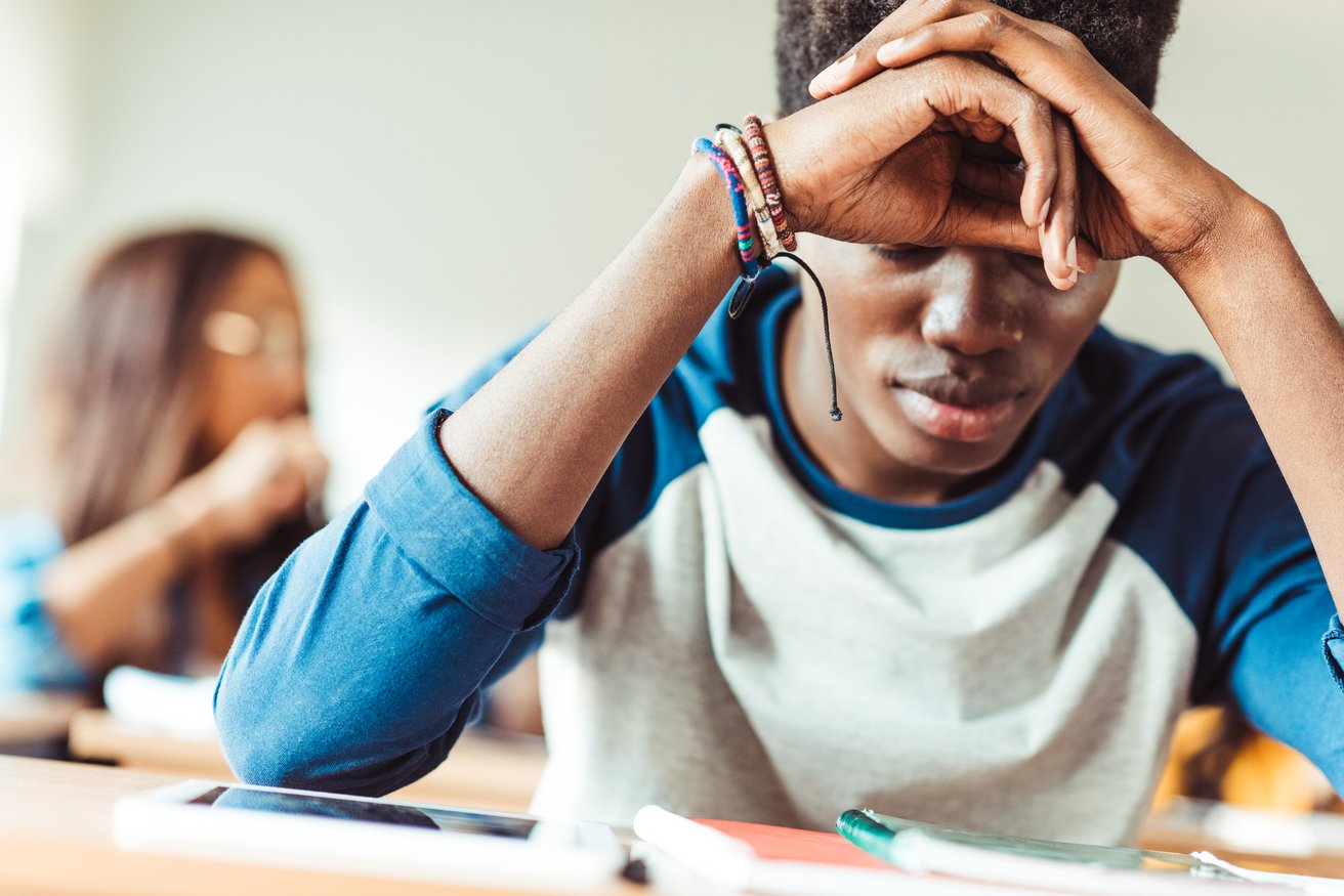 A teenager sits at a desk with their head in their hands, appearing distressed or overwhelmed in what looks like a classroom. Another student is visible in the blurred background.
