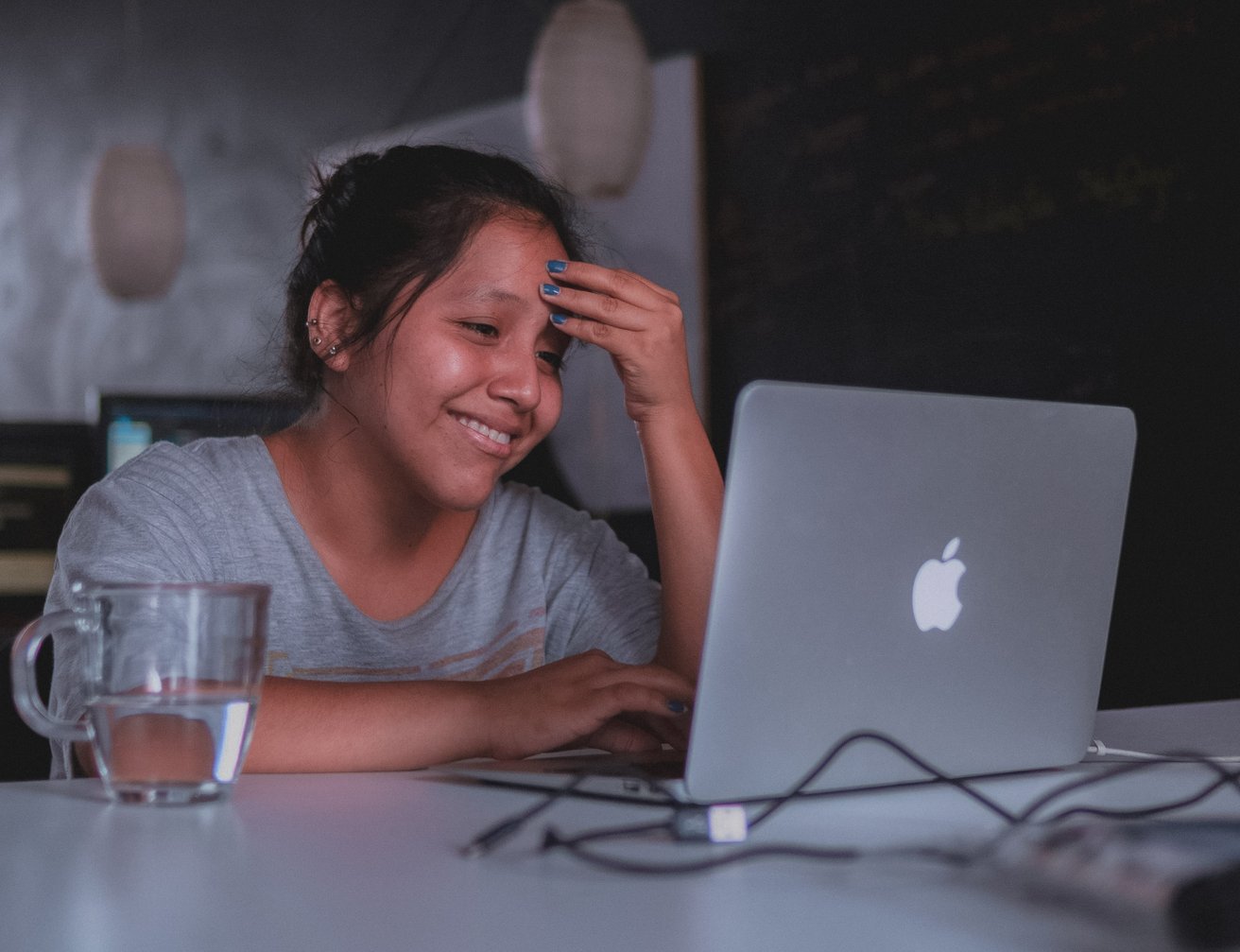A smiling teen sits at a table, looking happily at a laptop with one hand touching their forehead. A glass of water is visible next to them in an indoor setting.