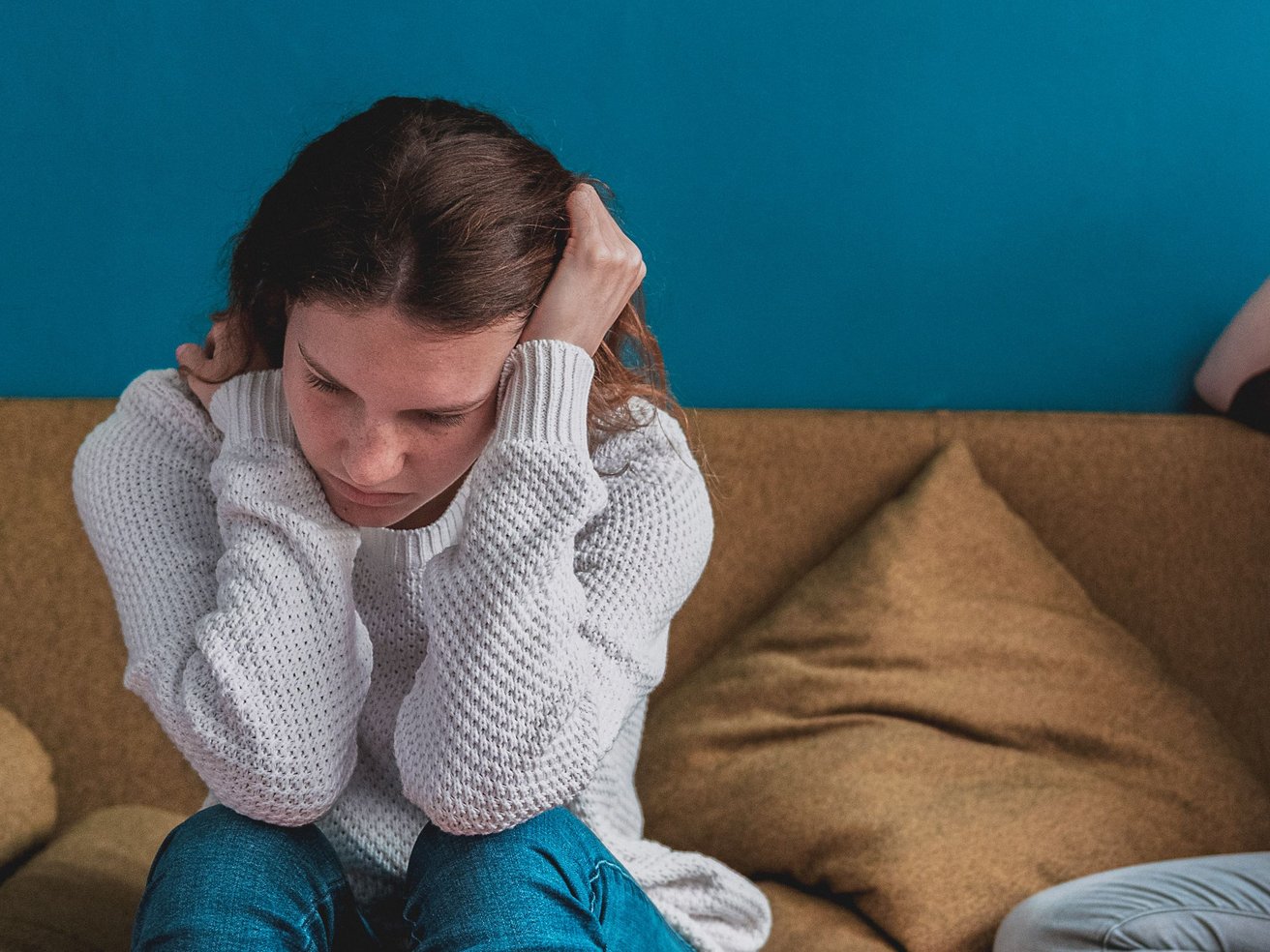 A young person, likely a teen, with brown hair sits on a brown sofa, looking down with a sad or distressed expression. One hand rests near their head, against a plain blue wall.
