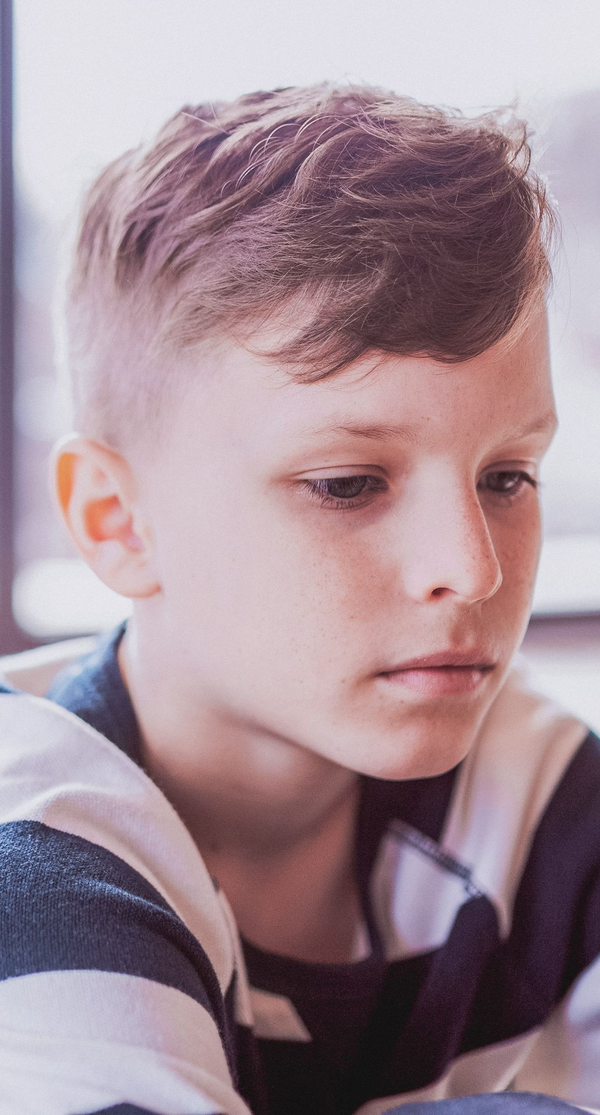 A close-up portrait of a young boy with short hair and freckles, looking down with a contemplative expression in a softly lit indoor setting. He appears to be deep in thought.