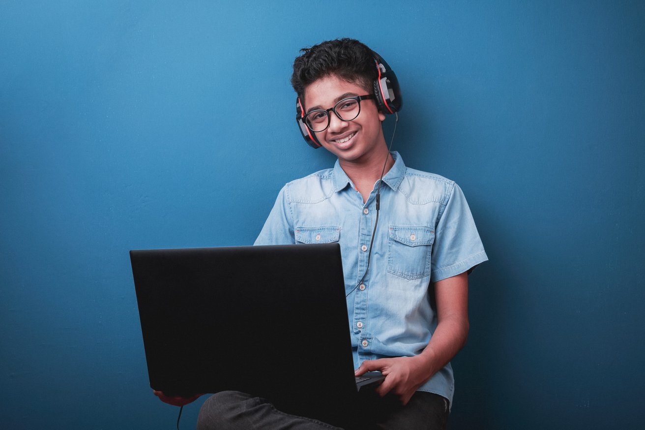A smiling South Asian teen boy, wearing glasses and headphones, holds an open black laptop, looking directly at the camera against a plain blue background. He appears happy and engaged.
