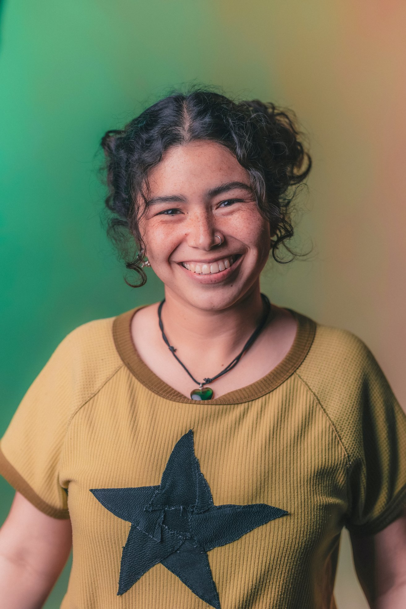 A smiling teen with curly hair and a nose piercing looks directly at the camera against a gradient green and orange background. They wear a ribbed shirt with a black star design, conveying a friendly and positive mood.