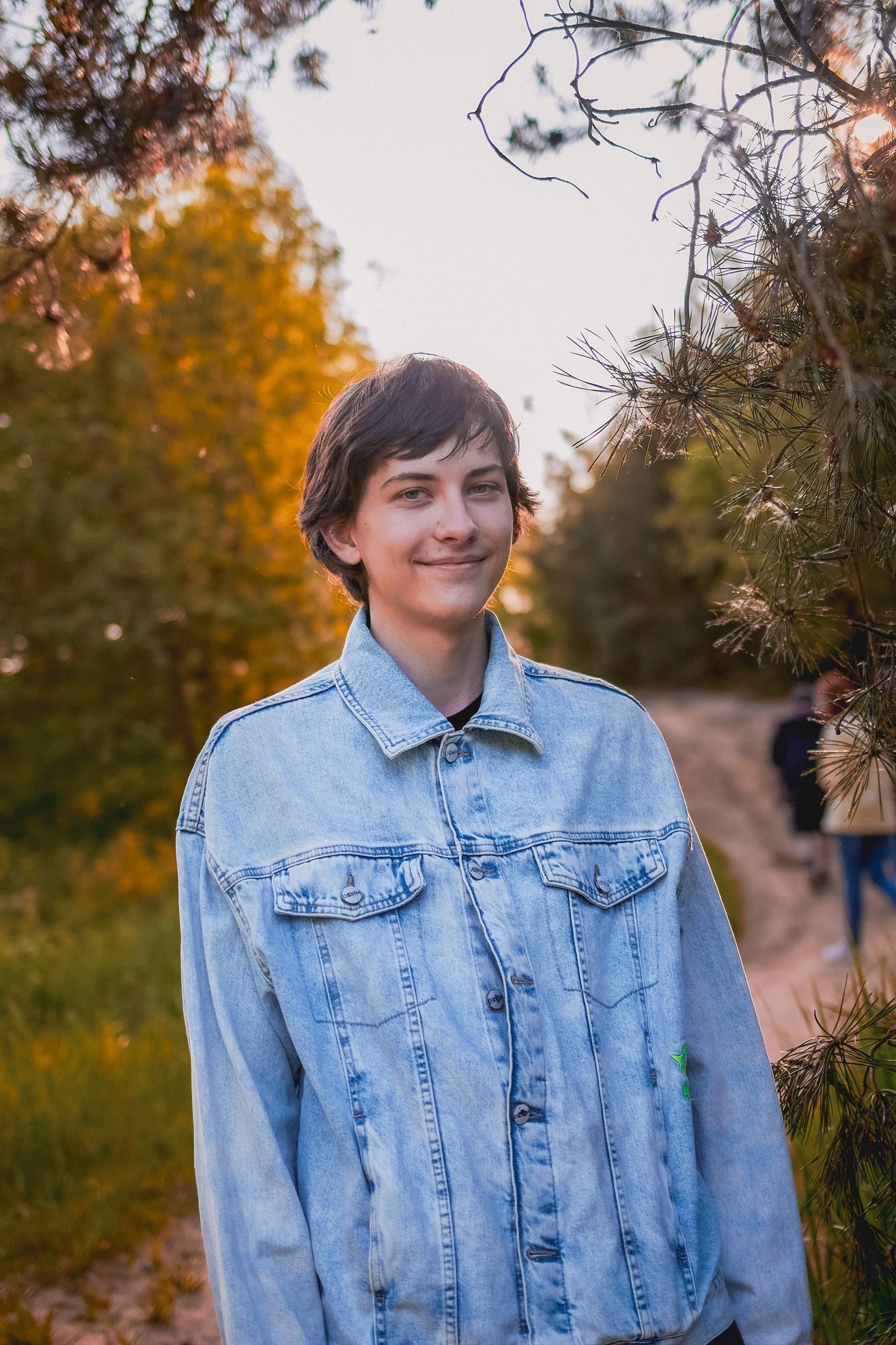 A smiling teenager wearing a denim jacket stands outdoors in a natural setting with golden sunlight filtering through trees. They look directly at the viewer with a friendly expression.