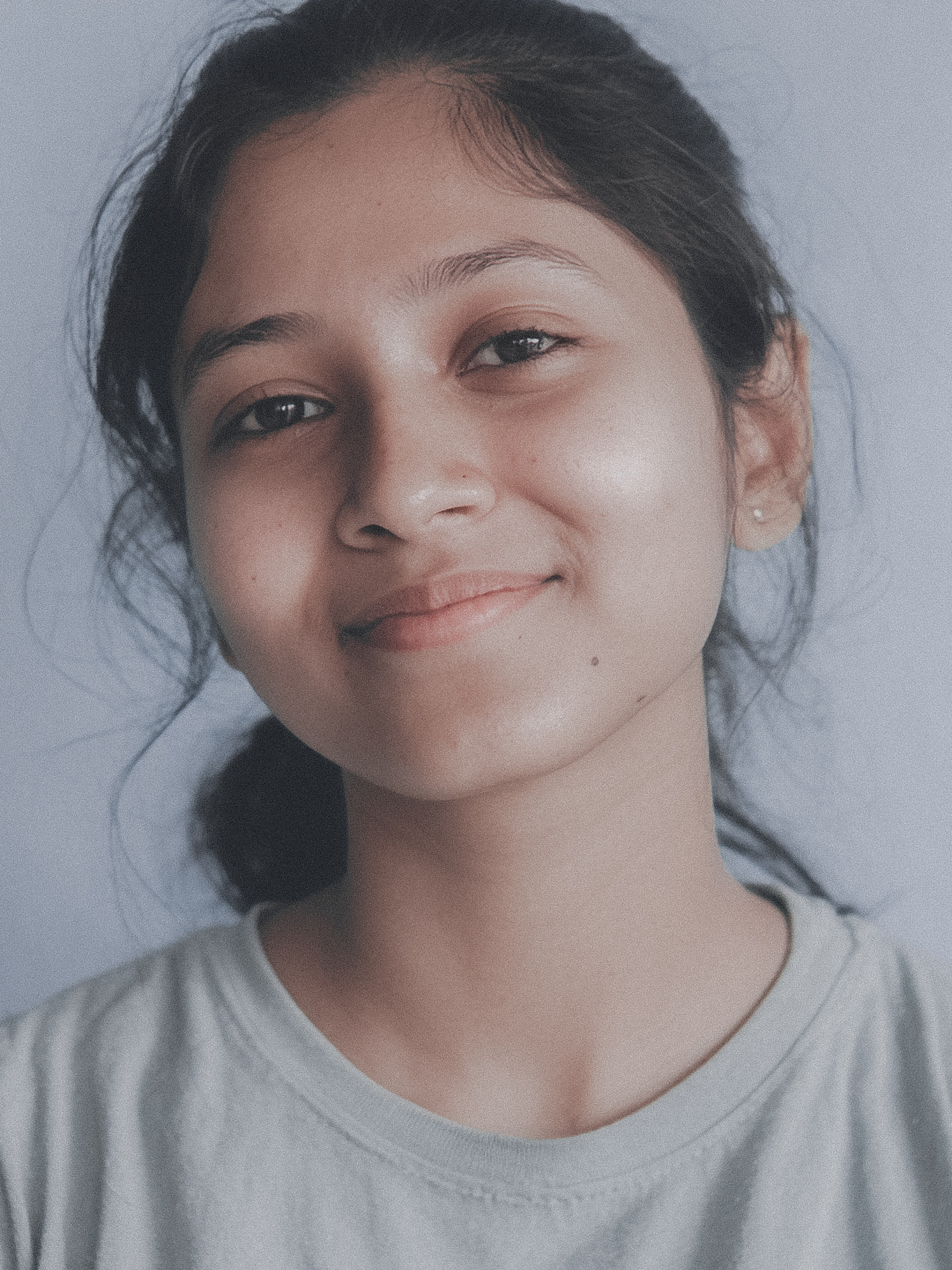 A young teenage girl with dark hair smiles gently, looking directly at the viewer in a close-up portrait against a light background. Her calm and friendly expression conveys approachability and warmth.