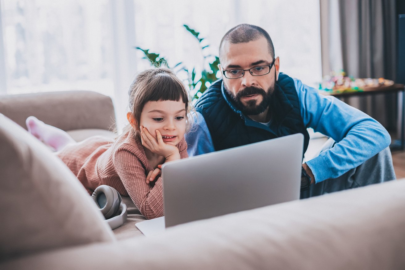 A young child and an adult man are intently focused on a laptop screen in a bright, cozy living room. The child lies comfortably on a sofa with headphones nearby, while the man sits on the floor beside her.