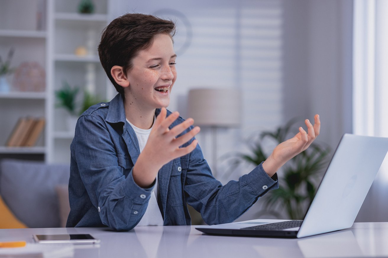 A happy teen boy with braces is smiling and gesturing towards a laptop screen, engaged in an online interaction from a bright home setting. He appears to be having a positive and engaging virtual session.
