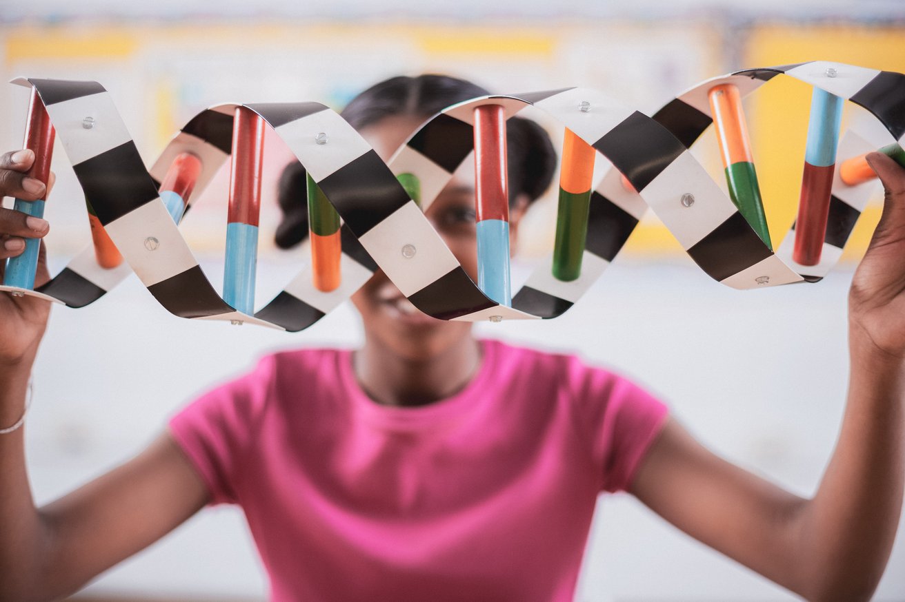 A young child holds a colorful DNA double helix model up to their face, peeking through it with a curious and engaging smile in a bright, indoor setting.
