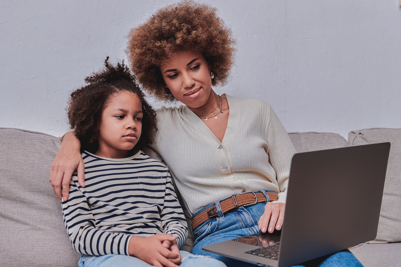 An adult woman with an arm around a child, both with curly hair, sitting on a sofa. The woman is looking at a laptop, while the child looks away with a pensive expression.