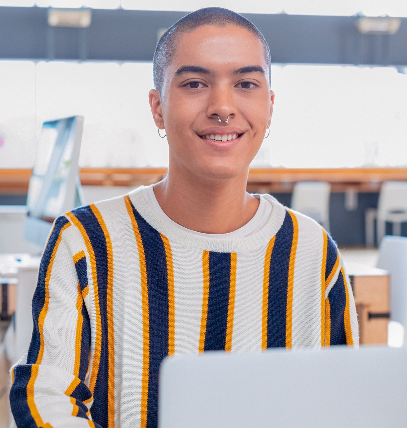 A smiling young adult with a shaved head and nose piercing looks directly at the camera while sitting at a desk with a laptop in a bright, modern indoor setting. They appear friendly and engaged.