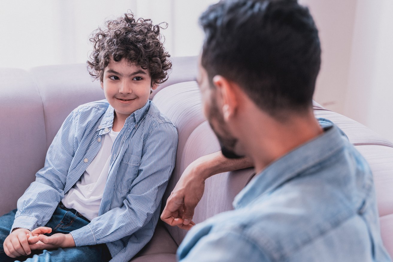 A young boy with curly hair sits on a light-colored sofa, looking up and smiling subtly at an adult. The adult, seen from behind, is leaning towards the boy, engaged in a calm conversation in an indoor setting.