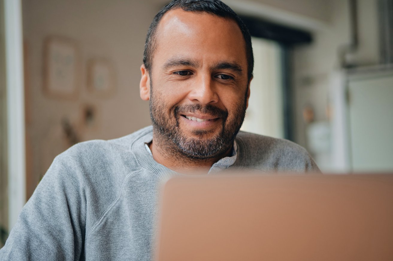 An adult man with a beard is smiling and looking down at a laptop screen in an indoor setting. He appears focused and engaged in an activity.