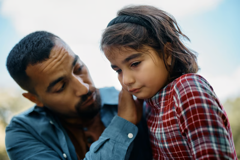 An adult gently comforts a young child who looks down with a solemn expression outdoors.