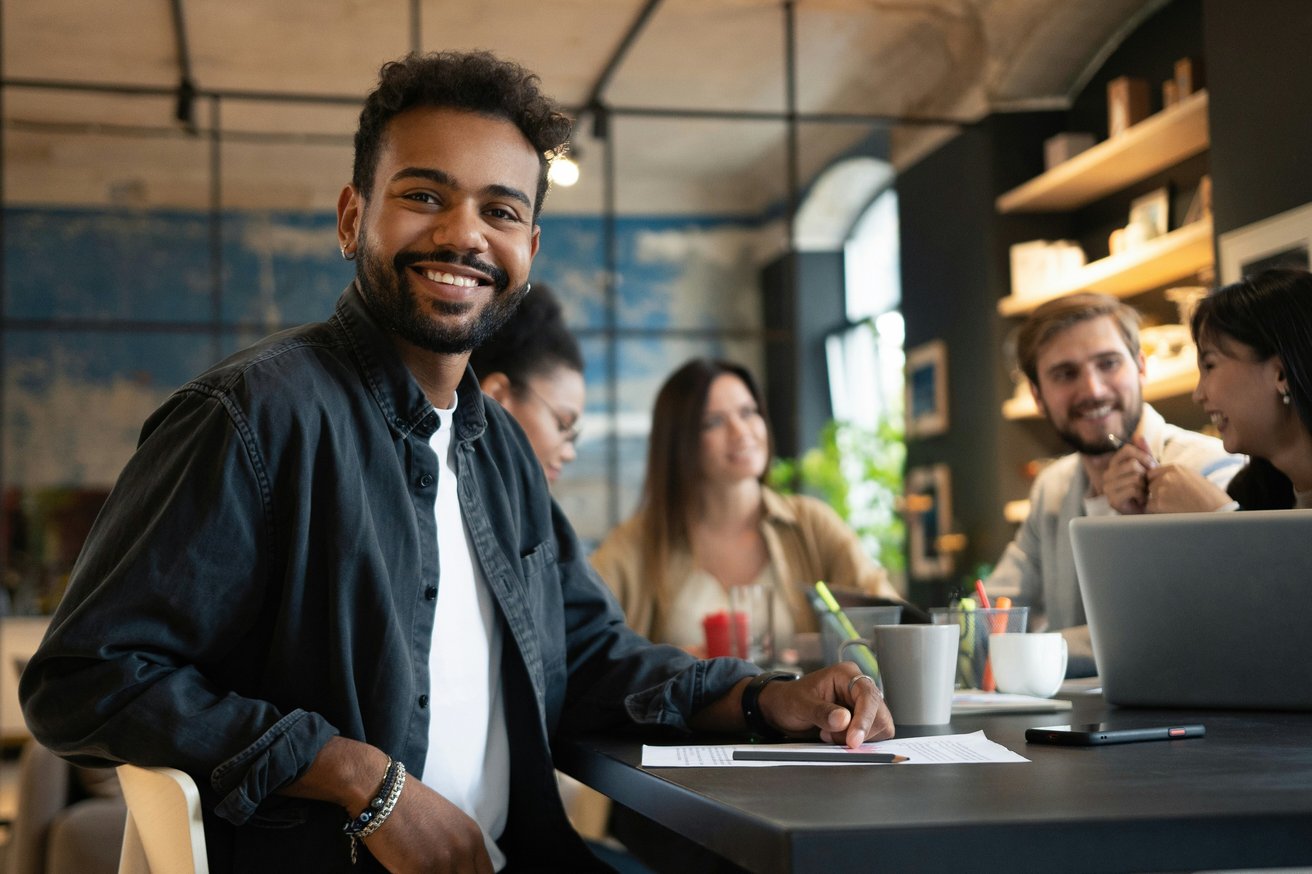 A smiling adult man with a beard looks directly at the camera while sitting at a dark table in a modern office space. Several other adults are blurred in the background, appearing to be engaged in conversation or working collaboratively.