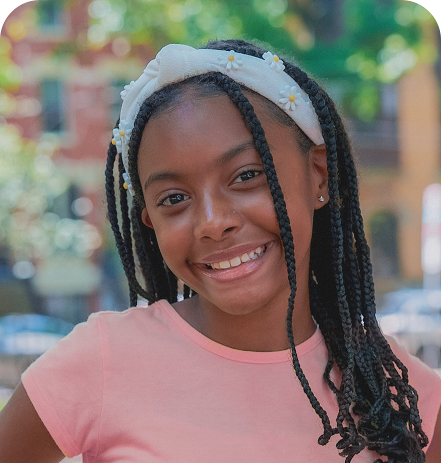 A young girl with long dark braided hair and a white daisy-decorated headband smiles brightly at the viewer. She is wearing a pink t-shirt and is captured in an outdoor setting with a blurred background.