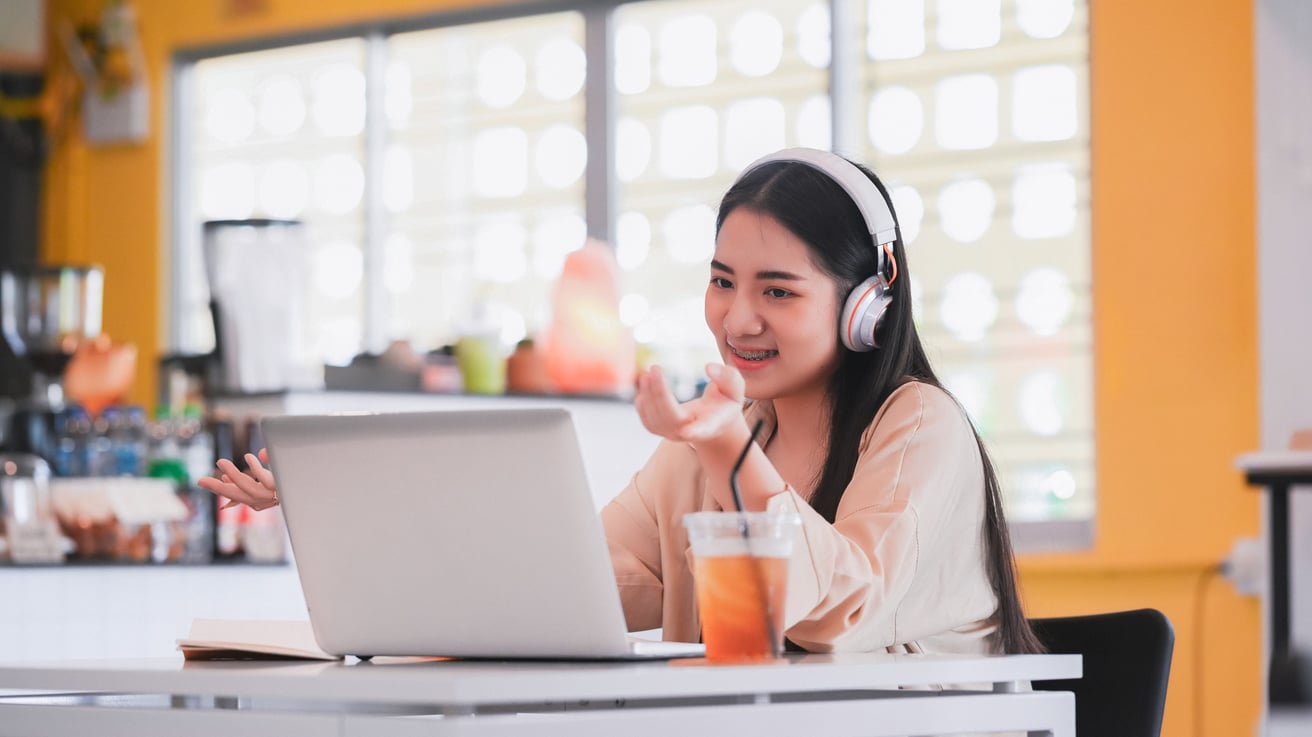 A smiling teen wearing headphones talks animatedly during a video call on a laptop in a bright, modern indoor setting. A drink sits on the table beside the laptop.