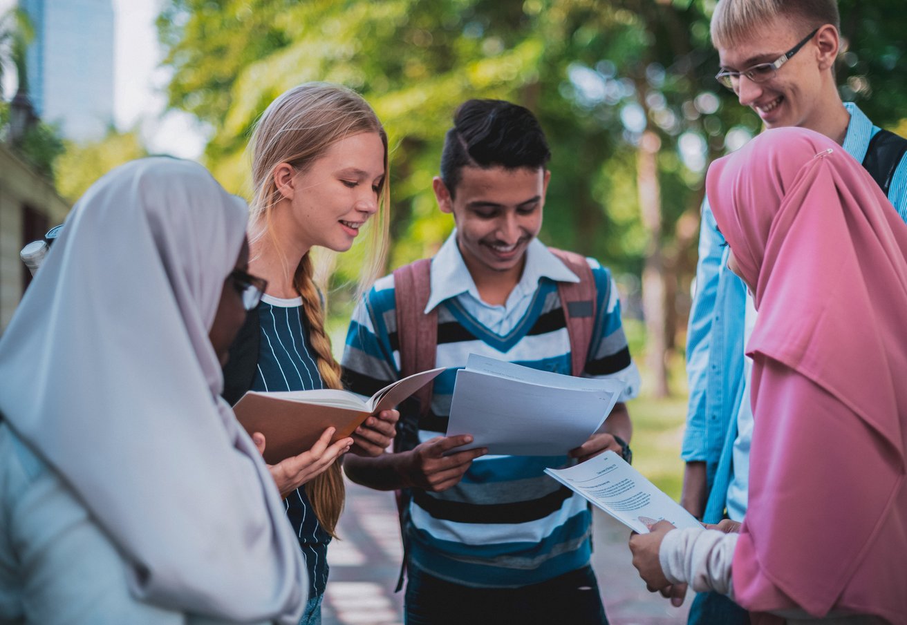 Five teenagers are gathered outdoors, smiling and looking at books and papers, engaged in a discussion or collaborative learning. The bright outdoor setting with green foliage suggests a positive and engaging environment.