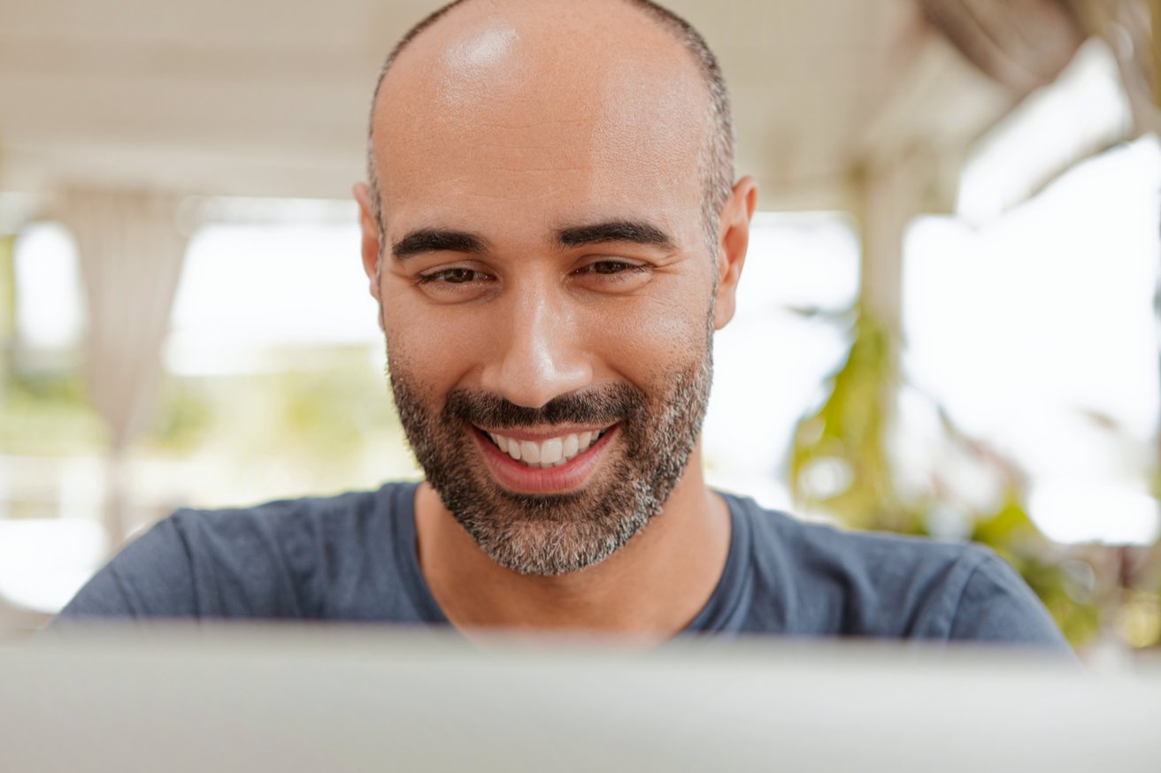 A smiling adult man with a beard looks down with a friendly and engaged expression. The background is softly blurred, suggesting a bright indoor or semi-outdoor setting.