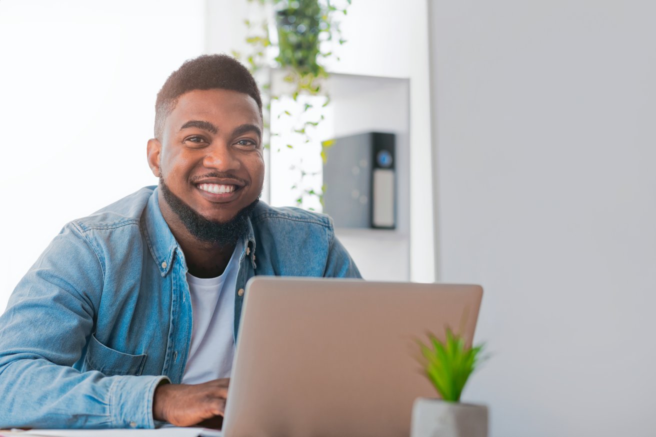 A smiling adult man sits at a desk with a laptop in a bright, modern office setting. He is looking directly at the camera with a friendly and approachable expression.