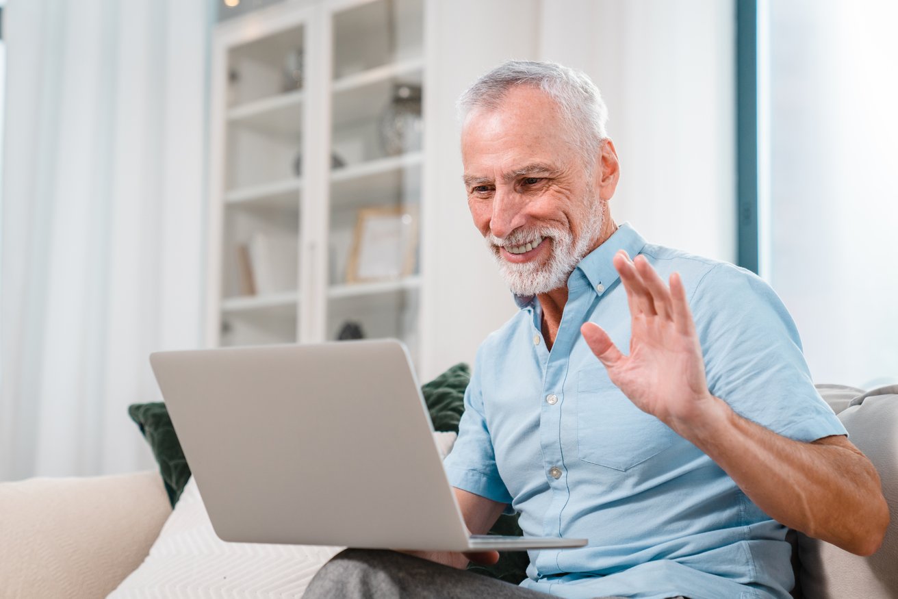 An older adult man with a gray beard smiles and waves at a laptop screen while sitting on a couch, engaged in a virtual conversation in a bright living room setting.
