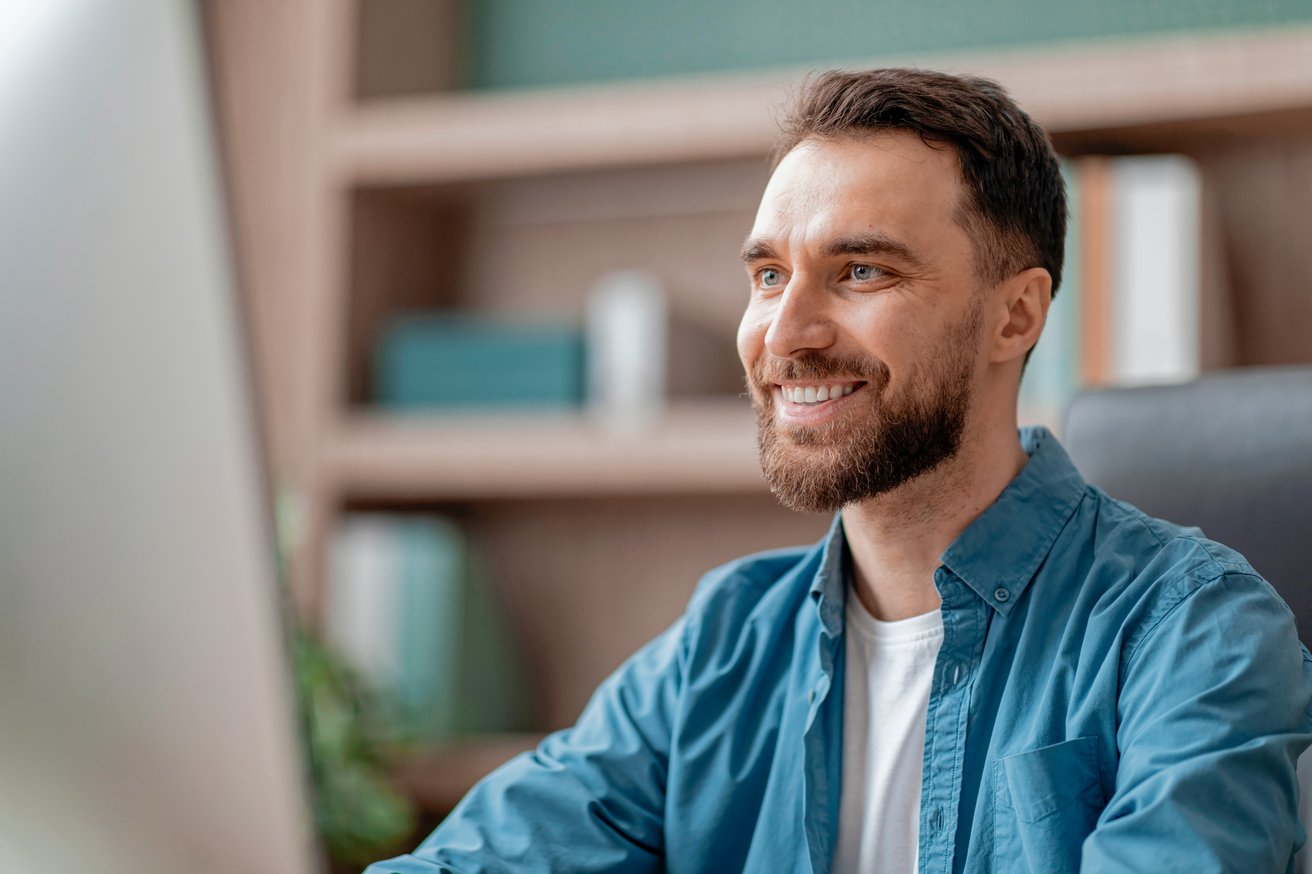 An adult man with a beard smiles genuinely while looking with interest at a screen in a bright indoor setting. Blurred shelves are visible in the background.