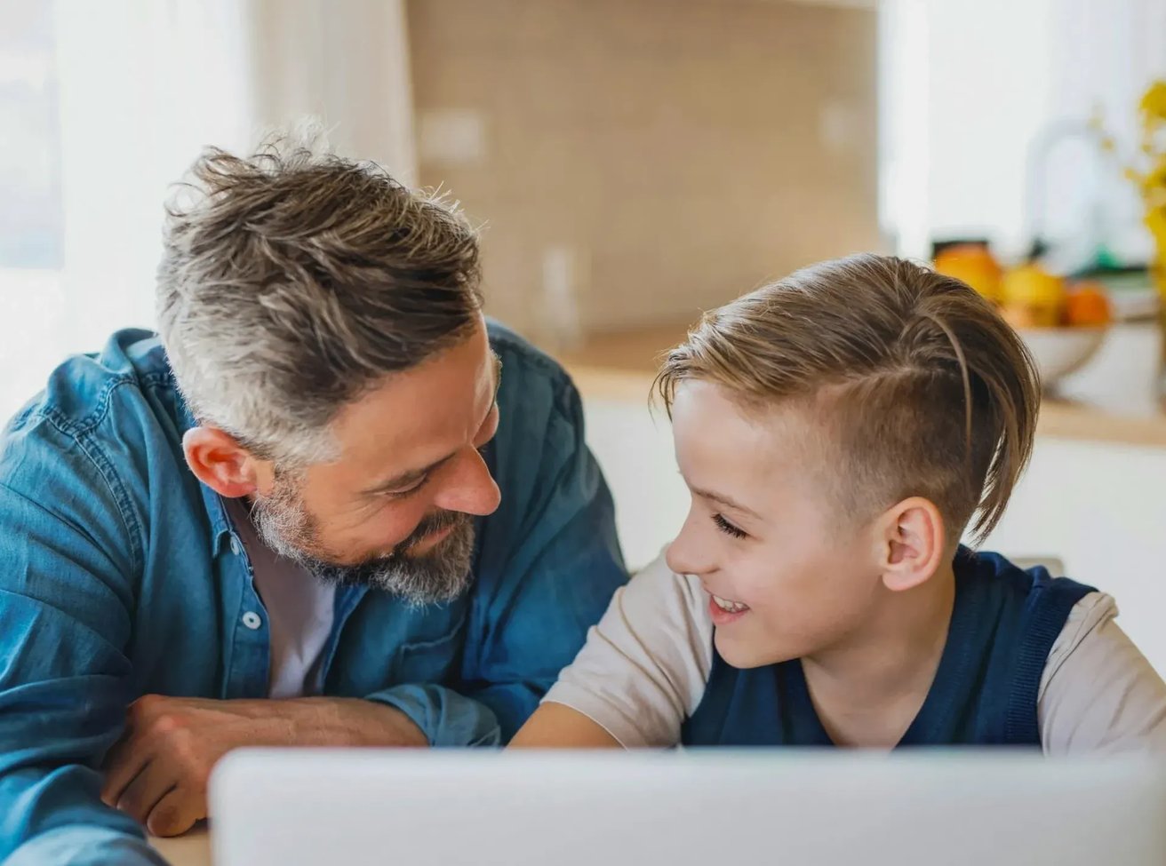 Dad and son in the kitchen smiling at each other with computer in front of them