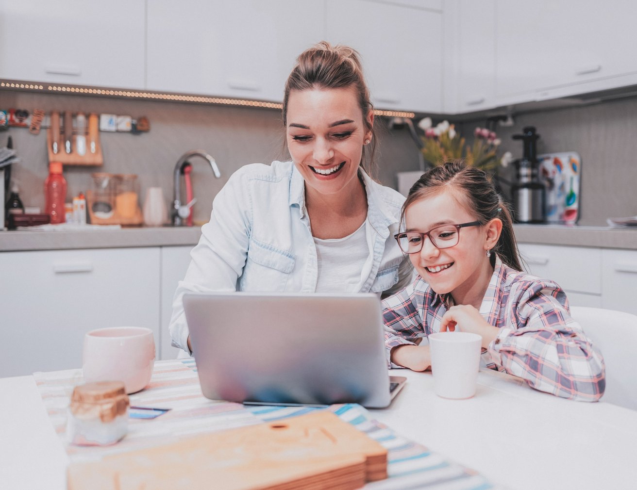 An adult woman and a young girl are happily looking at a laptop screen together at a kitchen table. They both smile as the woman puts her arm around the child, creating a warm, engaged atmosphere.