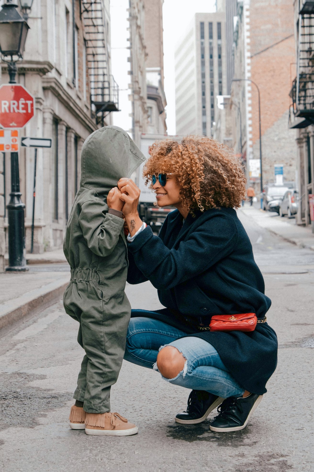 An adult with curly hair and sunglasses crouches down to hold the hands of a young child in a hooded jumpsuit on an urban street. The adult is smiling warmly at the child, conveying a sense of connection and care.