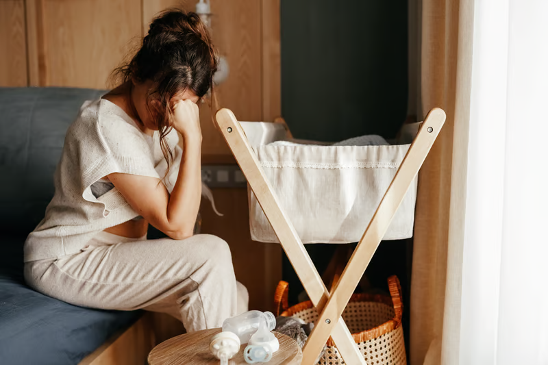 An adult sitting on the edge of a bed holding their head in stress next to an empty baby bassinet and feeding bottles.