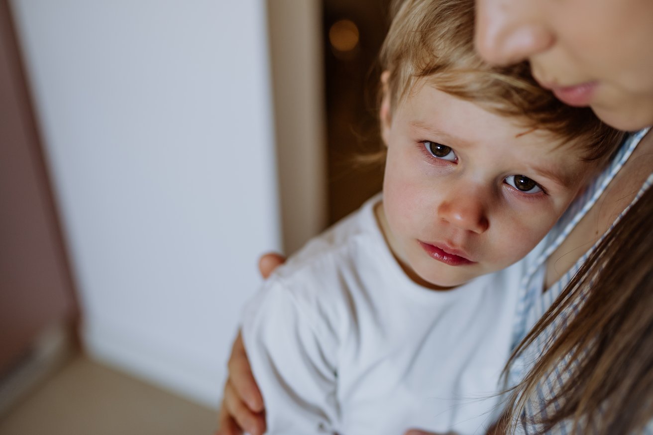 A young child, appearing sad with watery eyes, looks directly at the viewer while being gently held and comforted by an adult. The adult's chin and shoulder are visible, offering support in an indoor setting.