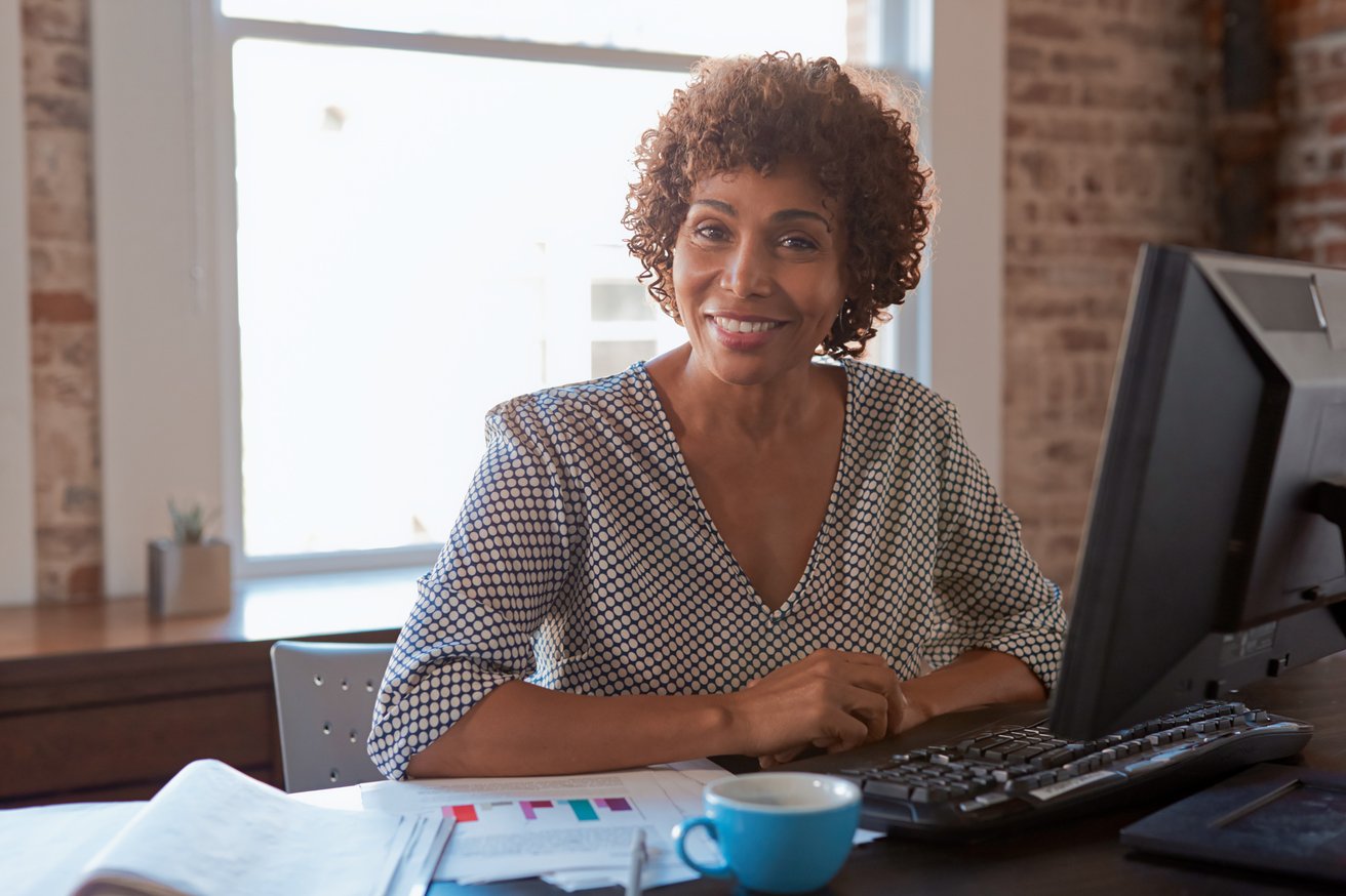 A smiling adult woman with curly hair sits at a desk, looking directly at the camera in a brightly lit office setting. A computer monitor, documents, and a coffee cup are visible on the desk, with a large window and brick wall in the background.