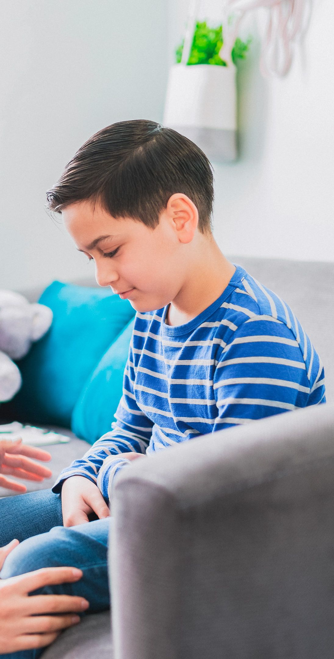A young boy wearing a blue and white striped shirt sits on a sofa, looking down intently at something in front of him. An adult's hands are gently placed on his leg, suggesting a supportive and comforting interaction in a relaxed indoor setting.