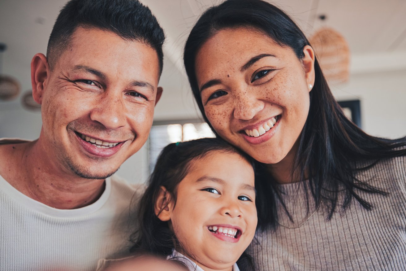 A close-up, smiling portrait of an adult man, an adult woman, and a young child taken indoors, conveying a sense of warmth and happiness among the family.