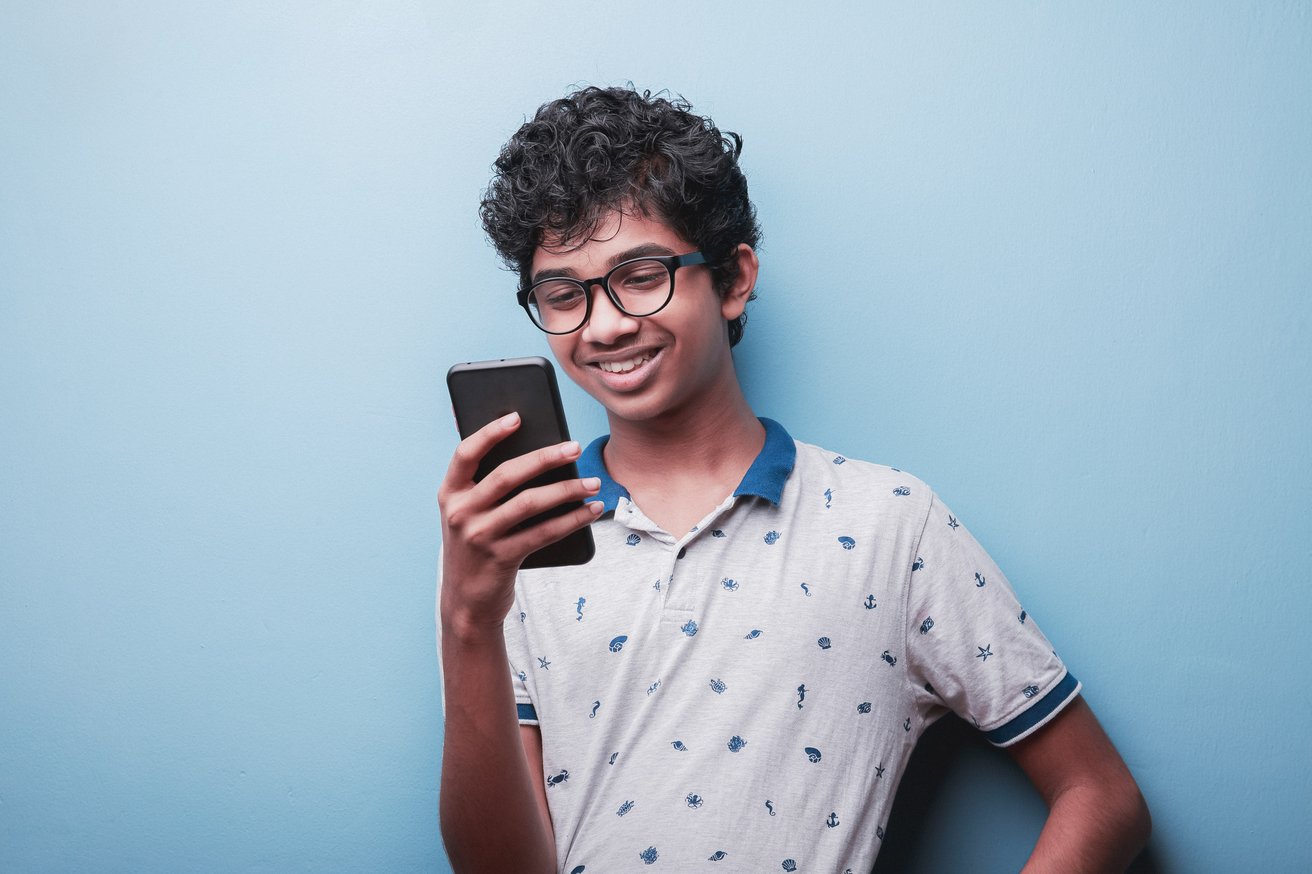 A smiling teenager with glasses holds and looks at a smartphone against a light blue background. The teen appears happy and engaged with the device.
