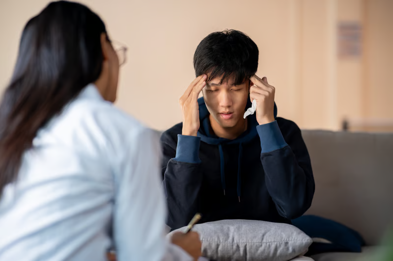 A distressed teen boy sits on a couch with his eyes closed and hands on his temples during a therapy session with a female professional.
