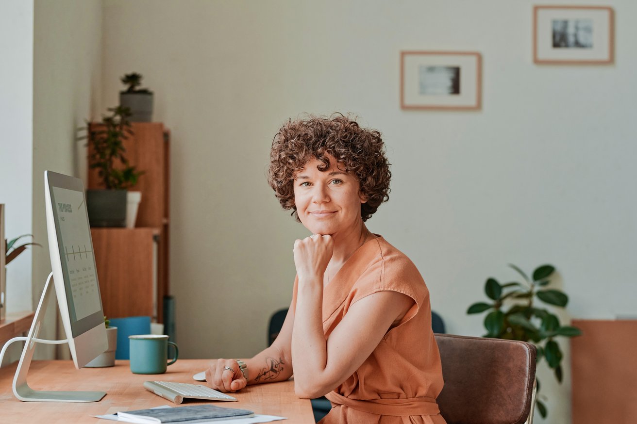 A smiling adult with curly hair sits at a modern desk in an office setting, looking directly at the viewer with a friendly expression. A computer monitor and keyboard are visible on the desk.