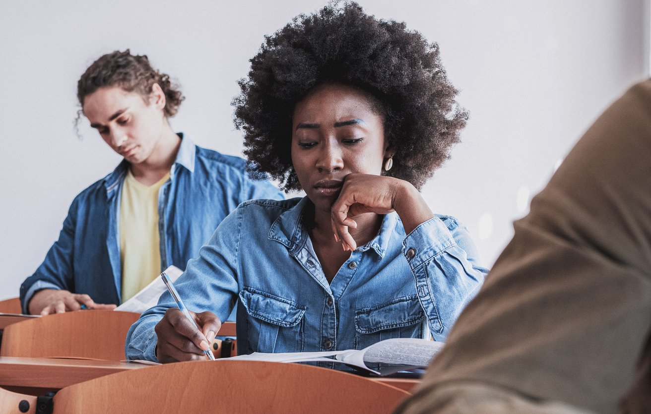 A young adult female student with dark curly hair is intently writing at a wooden desk in a classroom, appearing thoughtful. In the background, another young adult male student is also focused on his work.