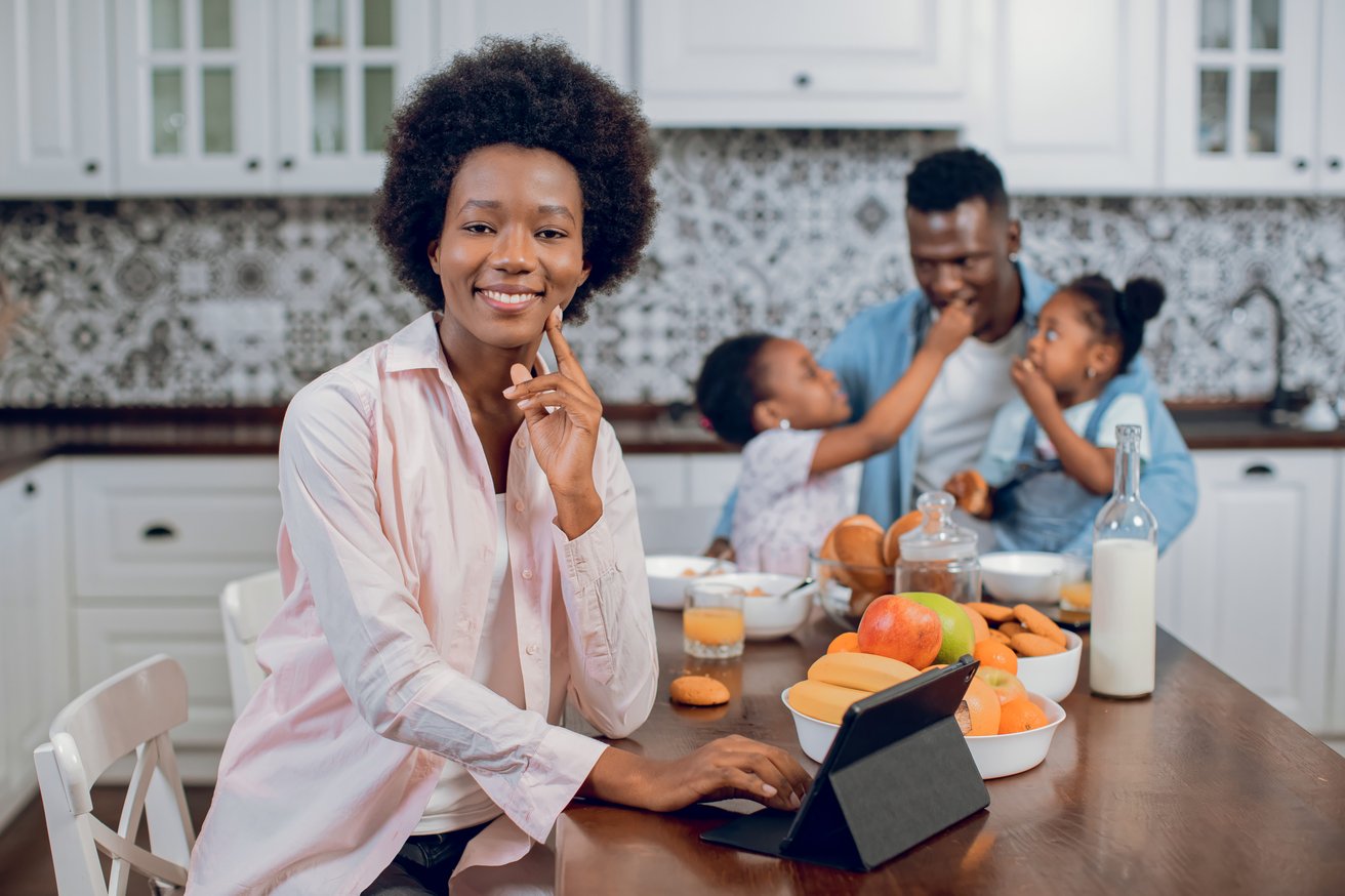 A smiling adult woman uses a tablet at a kitchen table, while in the blurred background, an adult man interacts playfully with two young children during a family meal.
