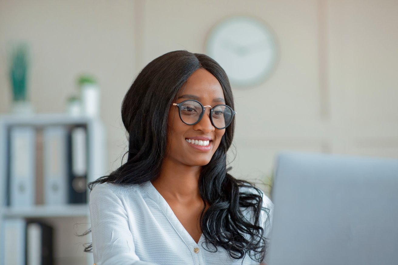 An adult woman with long dark hair and glasses is smiling warmly while looking attentively at a laptop screen. She appears to be in a professional office setting, conveying a friendly and engaged mood.