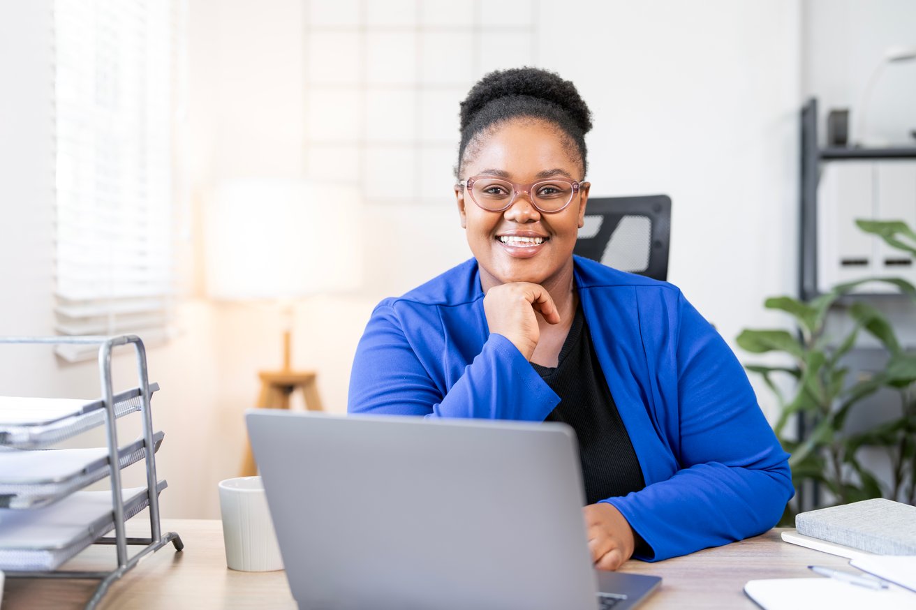 A smiling adult woman with glasses and a blue blazer is sitting at a desk, looking confidently at the camera. A laptop, coffee cup, and office supplies are visible on the desk, suggesting a professional work environment.