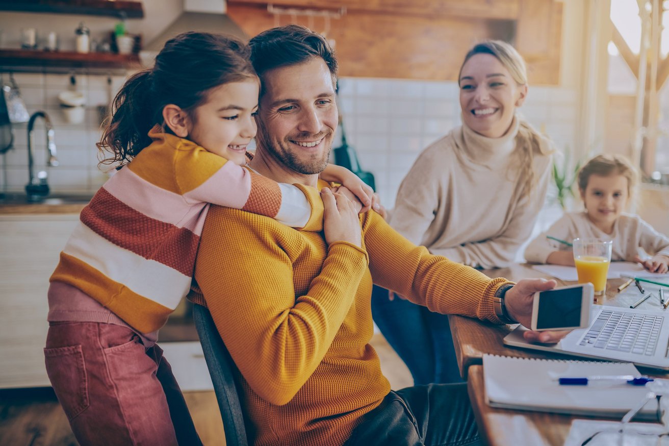 A happy family is gathered in a bright home setting. A young girl hugs an adult man who is holding a phone, while another young girl colors at the table, and an adult woman smiles in the background.