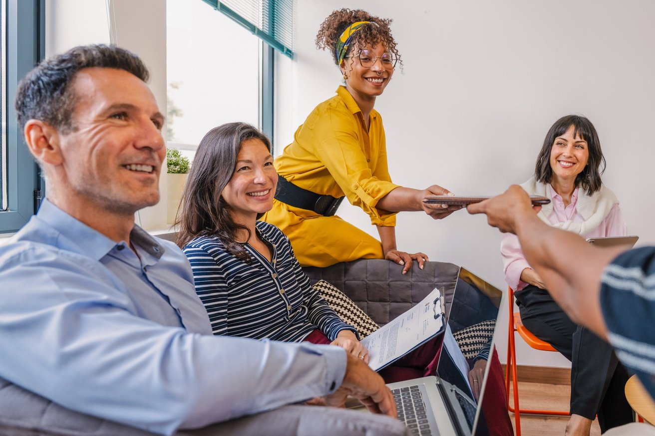 Four smiling adults are collaboratively working in a bright office space. One adult in yellow passes an item to another, while others engage with laptops and clipboards.
