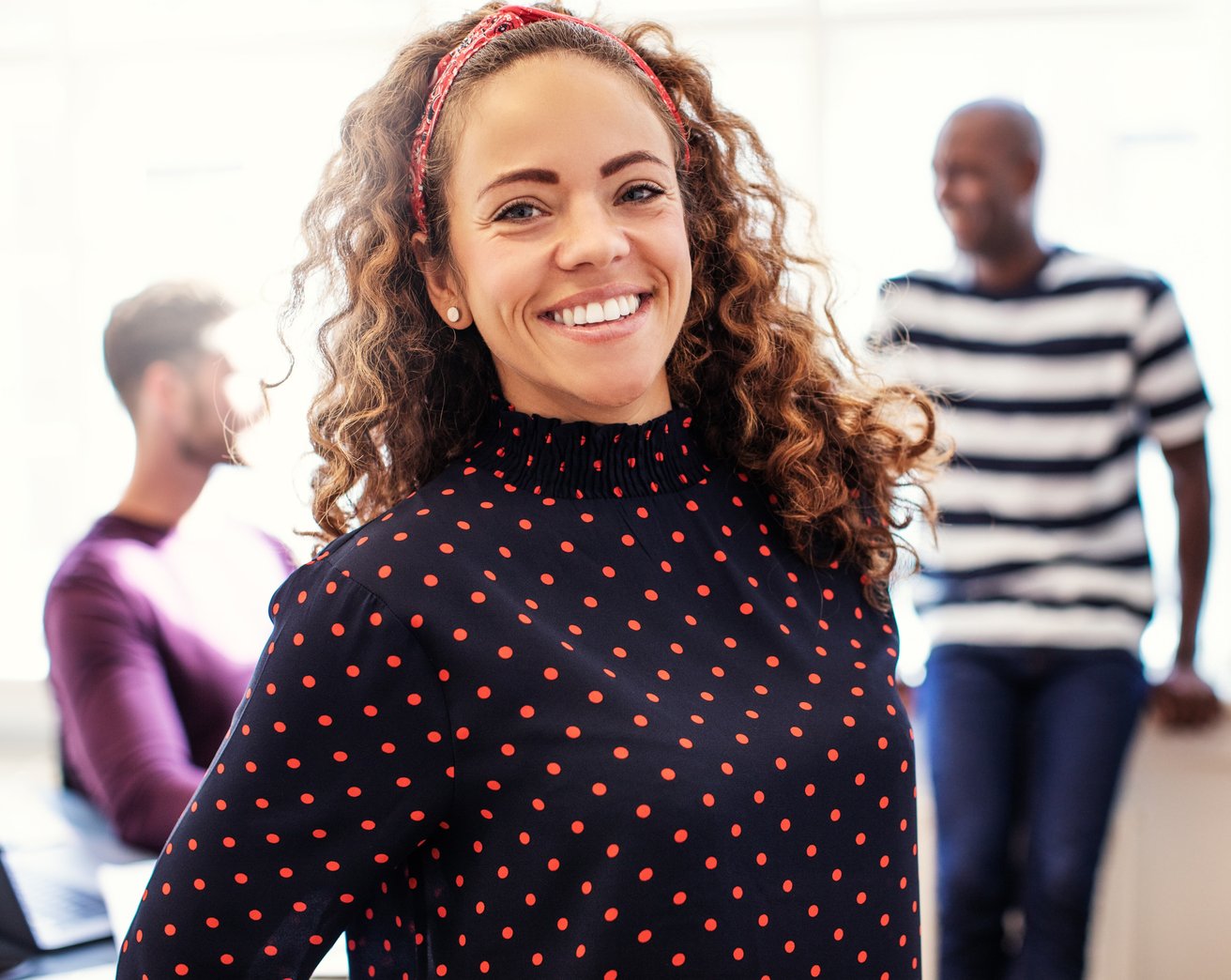 A smiling adult woman with curly hair and a red headband looks directly at the camera in a brightly lit office setting. Two blurred adult men are visible in the background, suggesting a collaborative environment.