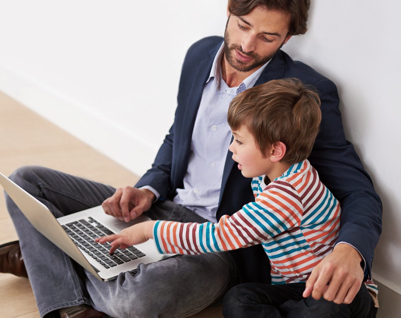 An adult man and a young boy are sitting on a light wood floor, leaning against a white wall, both looking at a laptop. The boy is pointing at the laptop while the man watches him attentively.
